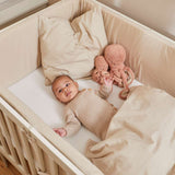 Bebé en una cuna FLEXA de color crema, de 140 x 70 cm, con una manta beige y un pulpo de peluche, en una foto tomada en una habitación infantil.