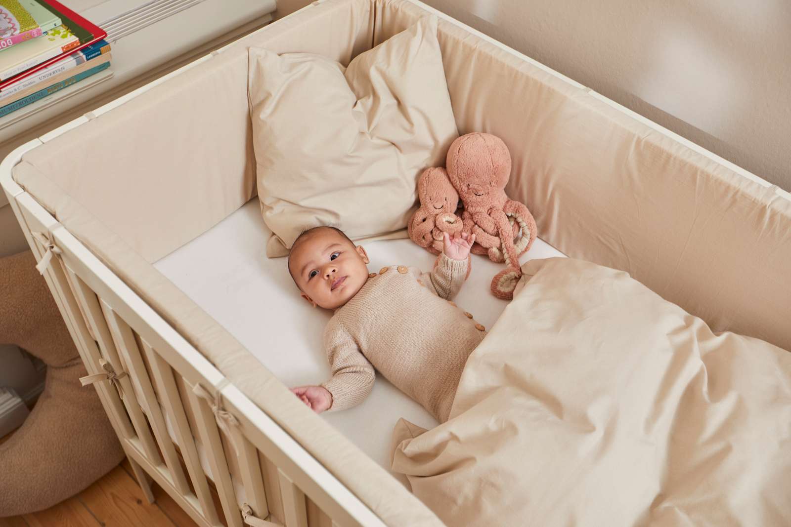 Bebé en una cuna FLEXA de color crema, de 140 x 70 cm, con una manta beige y un pulpo de peluche, en una foto tomada en una habitación infantil.