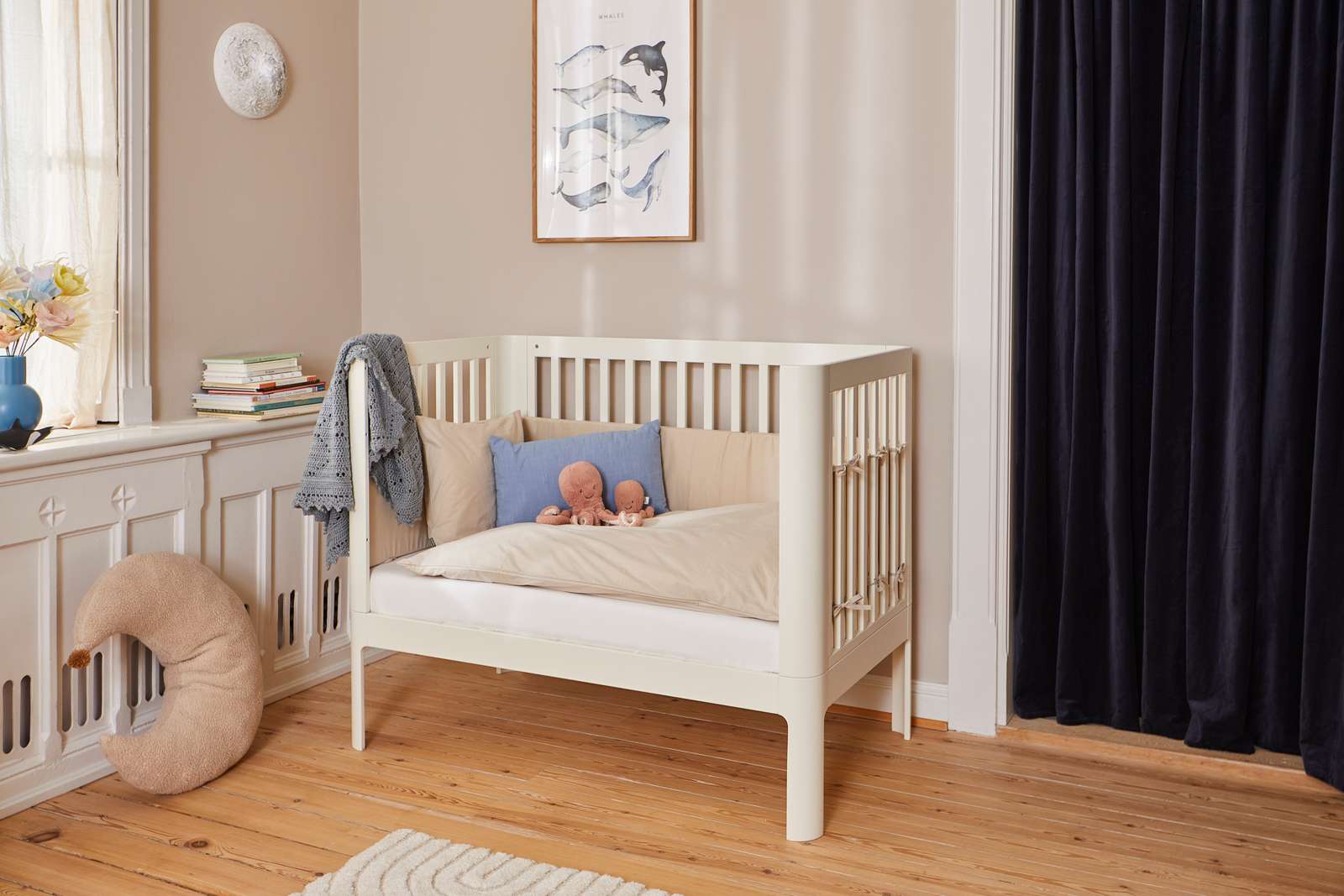 Cuna FLEXA color crema, 140 x 70 cm, de madera, con almohadas y juguetes, en una habitación infantil con iluminación suave y bordes redondeados seguros para los niños.