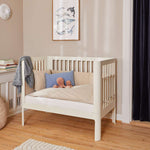 Cuna FLEXA color crema, 140 x 70 cm, de madera, con almohadas y juguetes, en una habitación infantil con iluminación suave y bordes redondeados seguros para los niños.