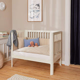 Cuna FLEXA color crema, 140 x 70 cm, de madera, con almohadas y juguetes, en una habitación infantil con iluminación suave y bordes redondeados seguros para los niños.