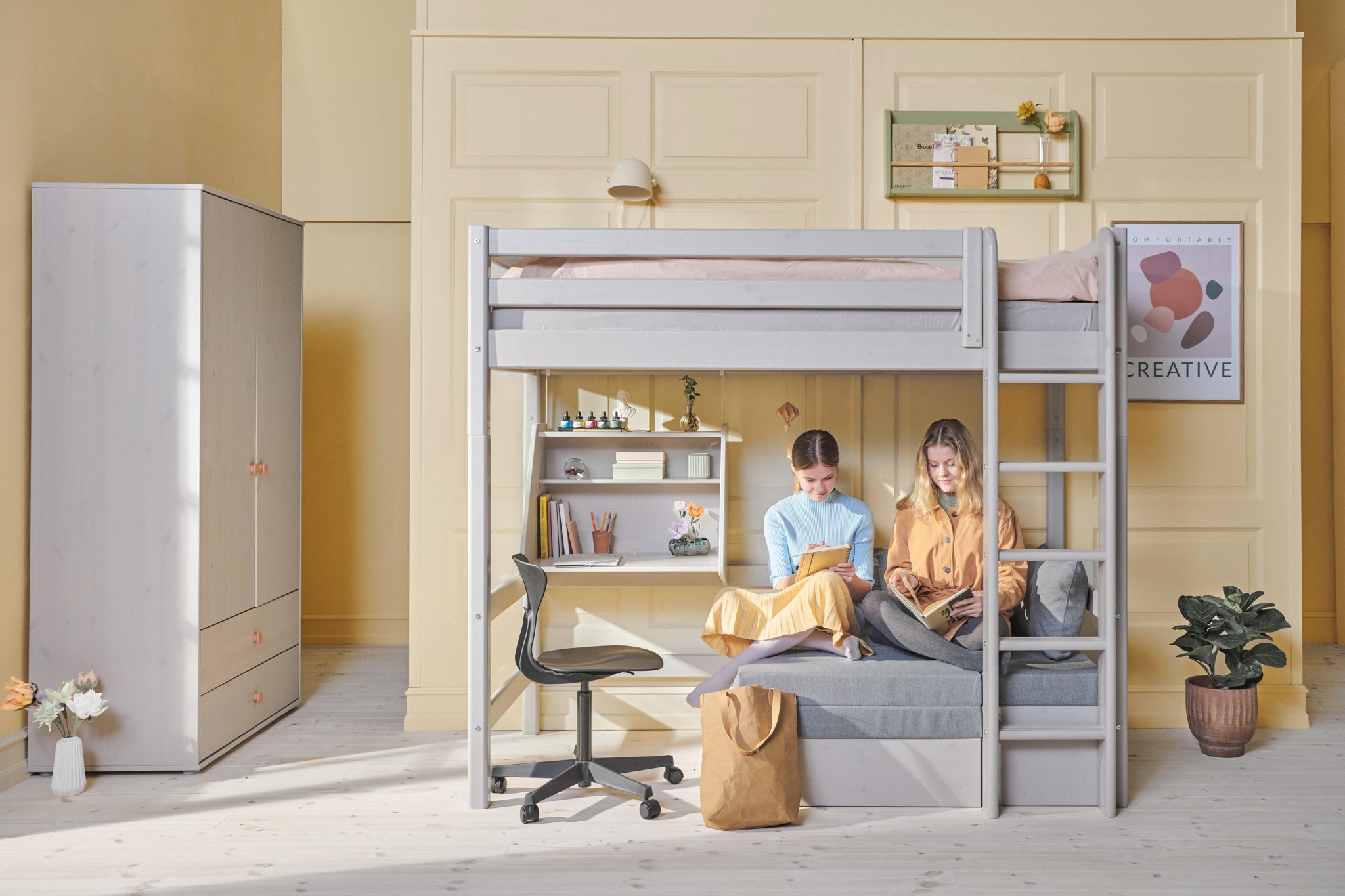 Cama alta FLEXA gris con sofá, escalera y barandilla de seguridad en una habitación luminosa con estanterías, plantas y cuadros en la pared.