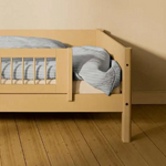 Barandilla de seguridad corta FLEXA SAND de WHITE & NOR en una cama infantil con ropa de cama a rayas azules en una habitación infantil de color beige.